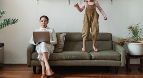 A woman working on her sofa and her child is next to her, jumping on the sofa