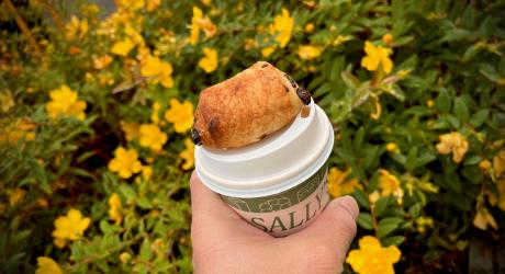 A hand holding a take away coffee cup with a croissant balanced on top with yellow flowers in the background.