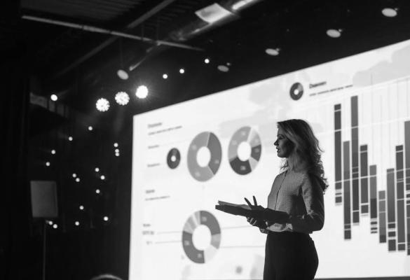 Woman giving a presentation with a large screen of statistics projected behind her.