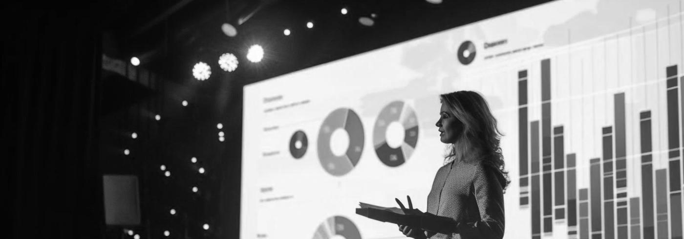 Woman giving a presentation with a large screen of statistics projected behind her.